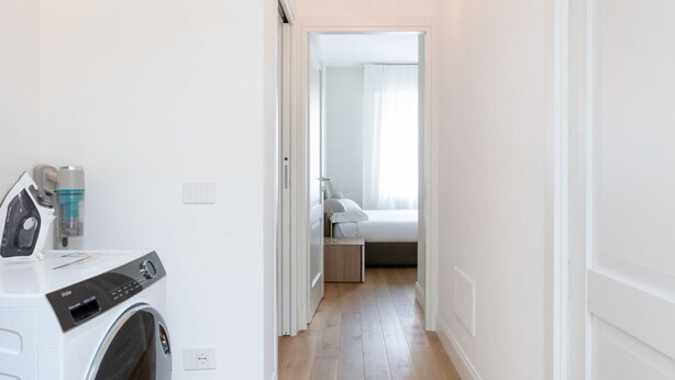 A laundry area with a front-loading washing machine, a bed with a nightstand, and a hallway leading to a bedroom.