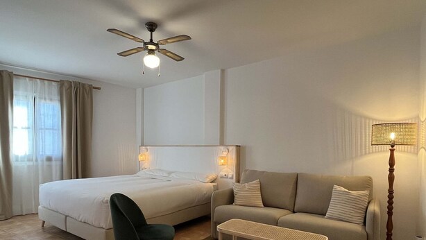 A hotel room with a bed, a sofa, a chair, a small table, a floor lamp, and a ceiling fan.