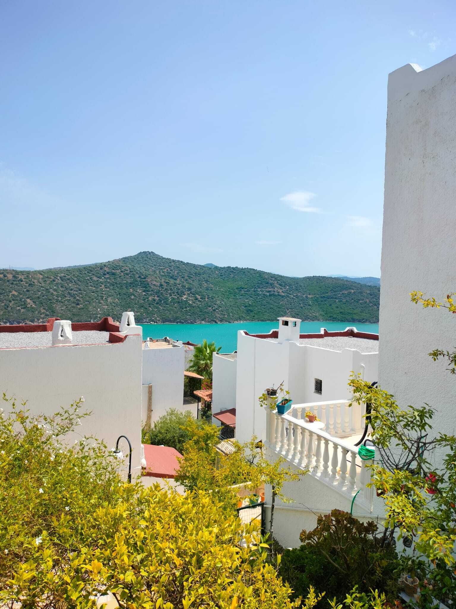 Zona residencial costera con edificios blancos, vista al mar y colinas verdes al fondo.