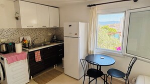 A compact kitchen with white cabinets, a refrigerator, a small dining table with two chairs, and a window with a view of greenery. - Peaceful Studio by the Beach in Bodrum (Muğla)