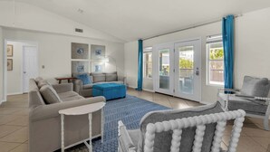 Ein geräumiges Wohnzimmer mit einer Couch, einem Hocker und einem Sessel, einem Deckenventilator und Blick durch Glastüren auf einen Außenbereich.