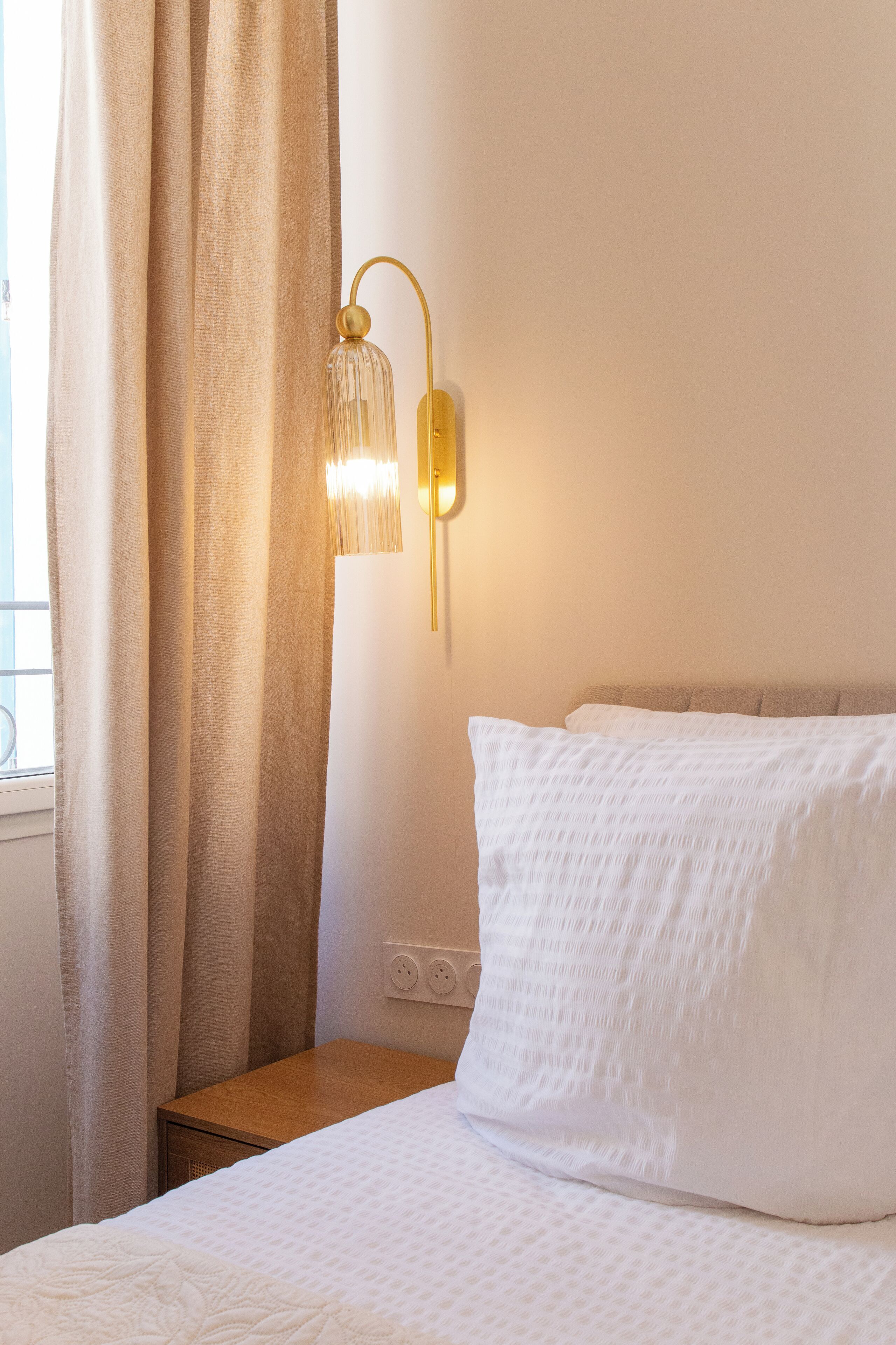Una cama bien hecha con ropa de cama blanca, una mesita de noche de madera, una lámpara de pared y una ventana con cortinas.
