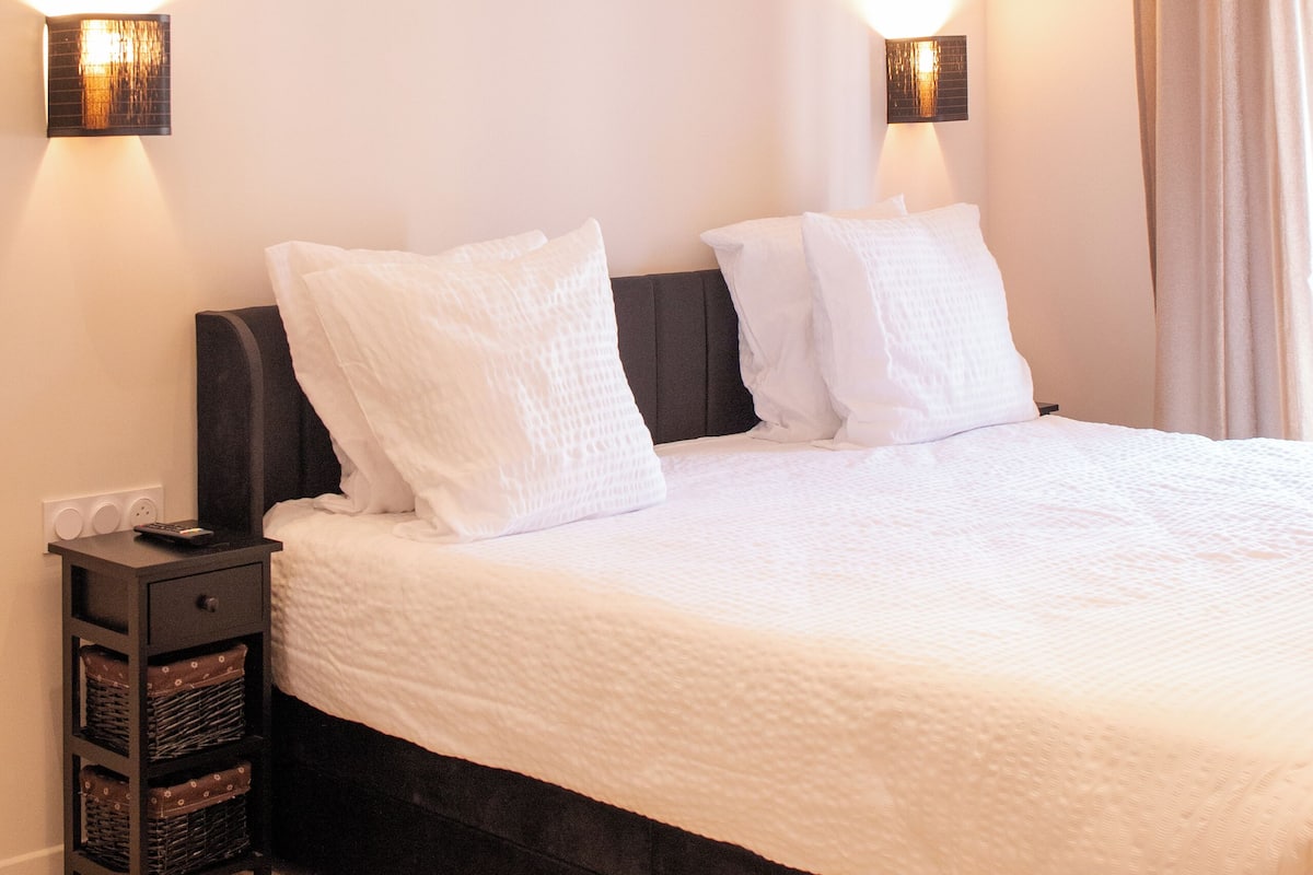 A neatly made bed with white bedding, a dark headboard, and two wall-mounted light fixtures on either side.