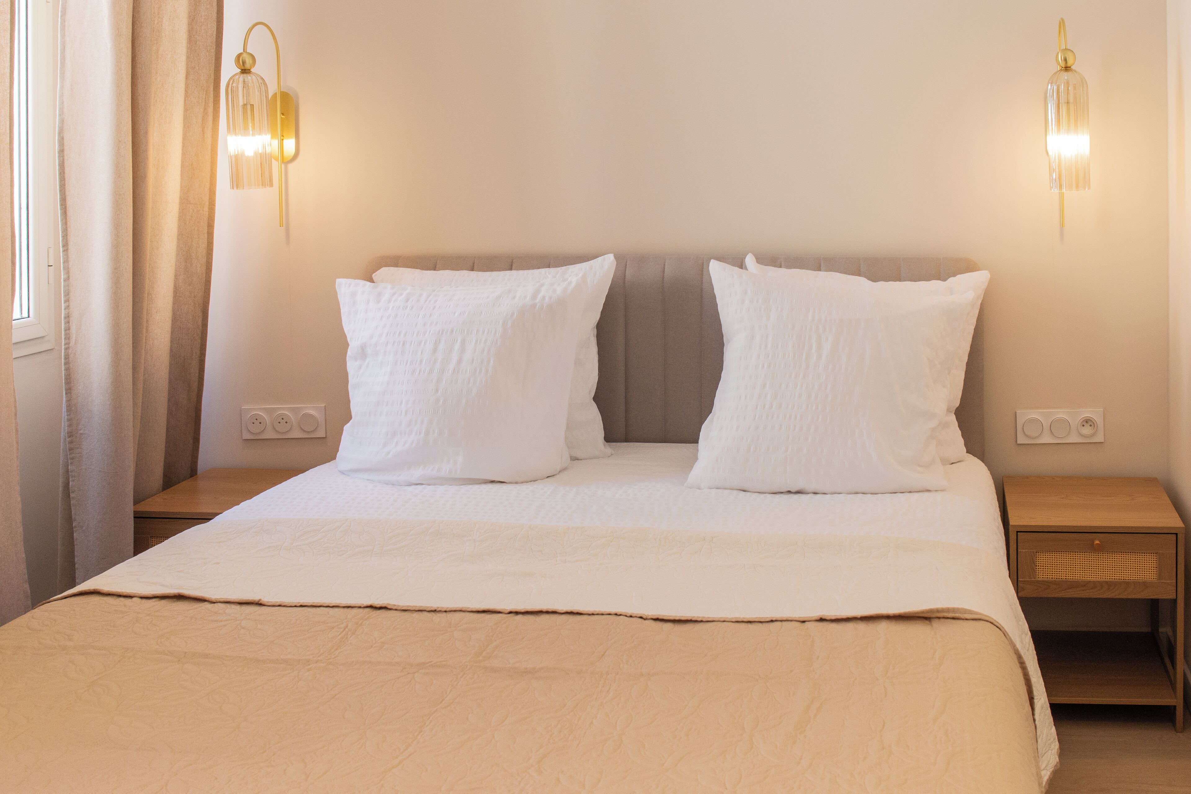 A neatly made bed with a beige bedspread and two white pillows, a wooden nightstand with a woven basket, and two wall-mounted light fixtures.