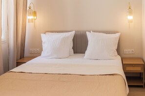 Uma cama bem arrumada com colcha bege e duas almofadas brancas, uma mesinha de cabeceira de madeira com cesto de vime e duas luminárias fixadas na parede.