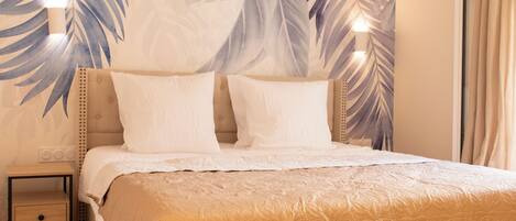 A neatly made bed with a beige quilt, a nightstand with a drawer, and a wall with a tropical leaf pattern.