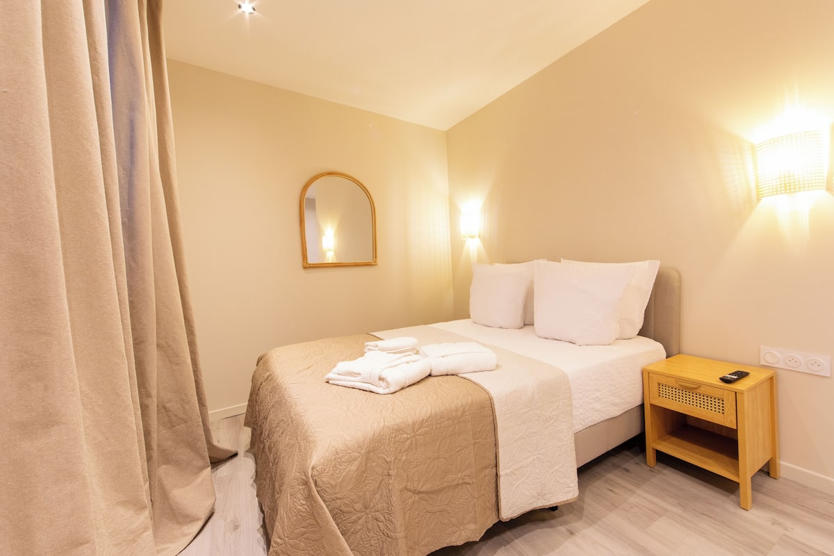 A neatly made bed with a beige bedspread, a bedside table with a lamp, and a wall-mounted mirror.