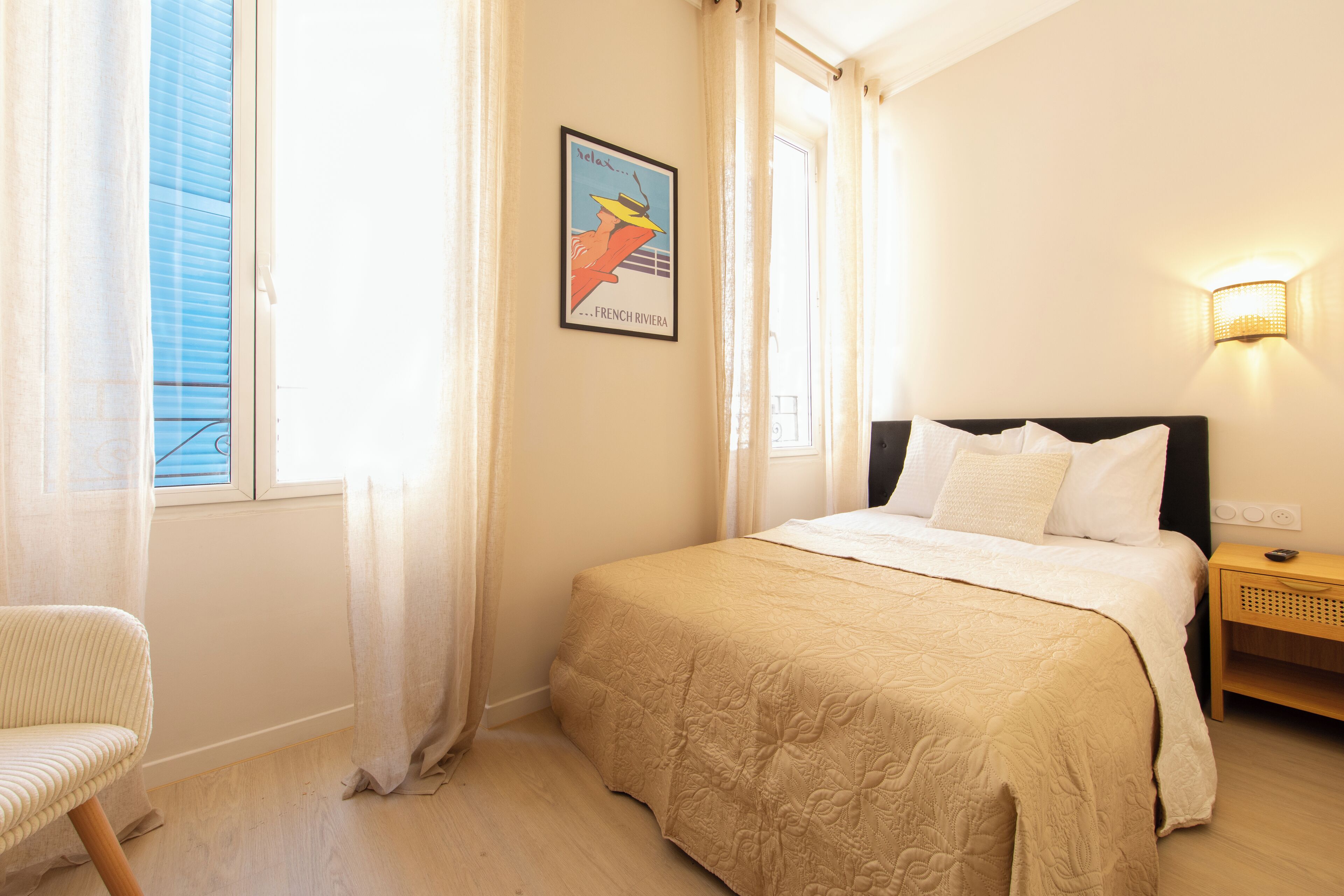 A bedroom with a bed, a chair, a nightstand, a wall-mounted lamp, and a window with curtains.