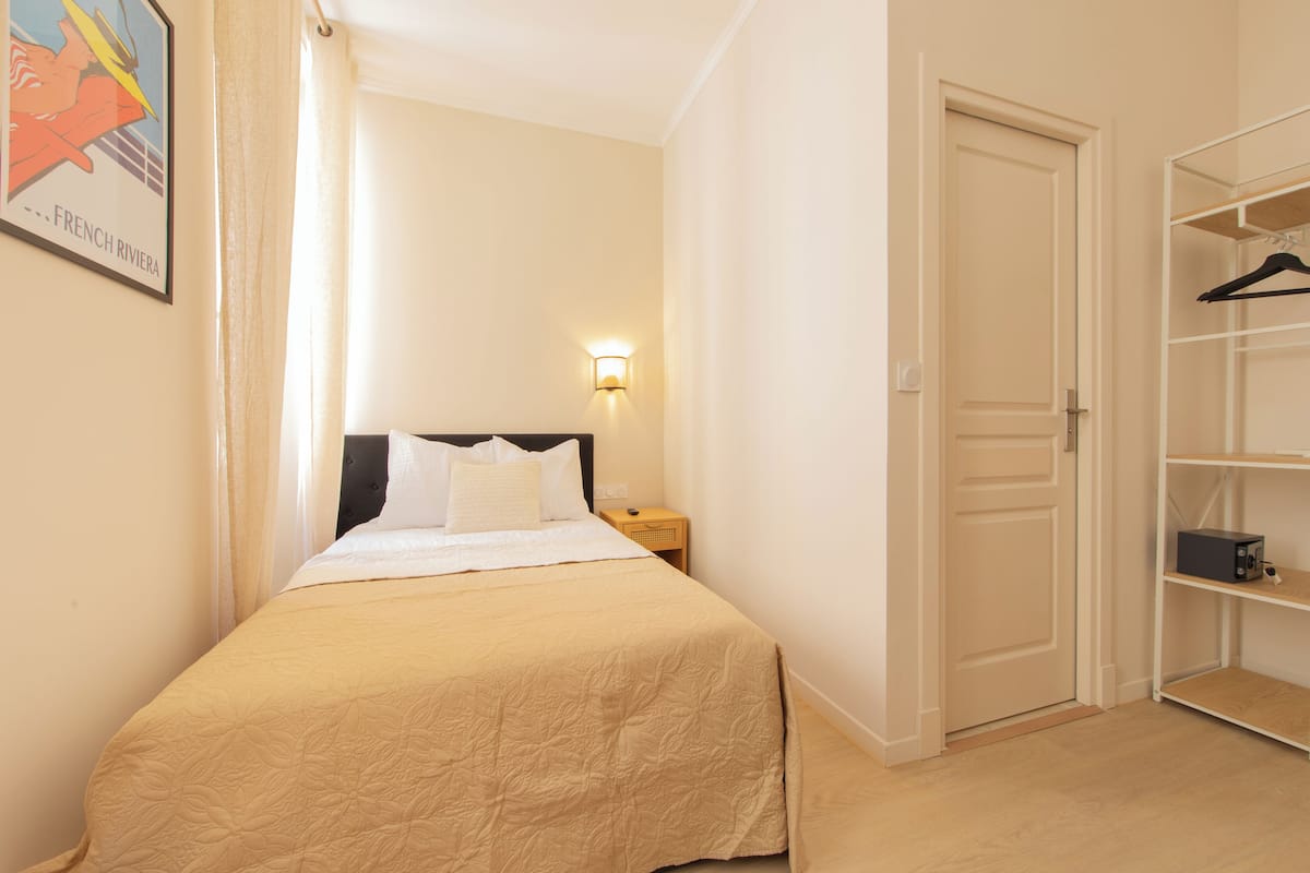 A bedroom with a bed, a nightstand, a wall-mounted light, a door, and a closet with shelves.