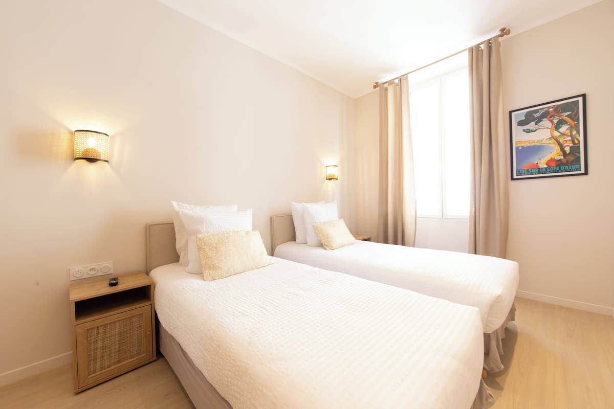 A hotel room with two beds, a nightstand, wall-mounted lights, a window with curtains, and a poster on the wall.