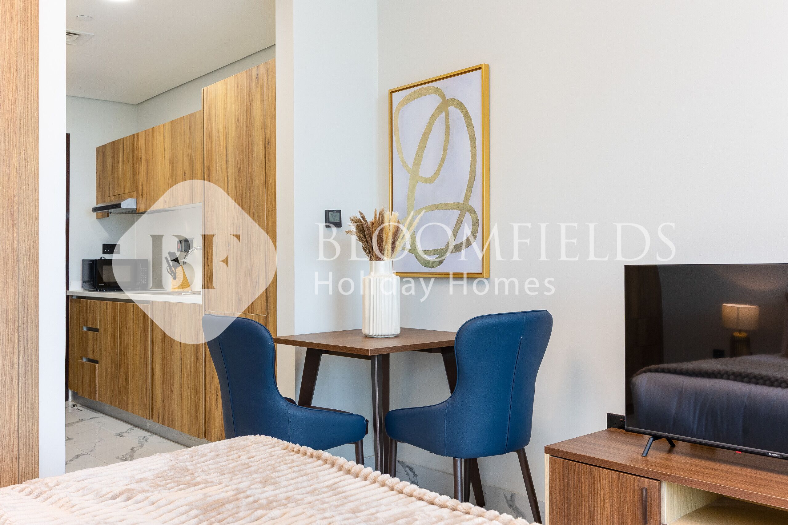 Ein modernes Hotelzimmer mit einem Bett, einem Fernseher, einem Schreibtisch mit zwei Stühlen, einer Küchenzeile und einem gerahmten Kunstwerk an der Wand.