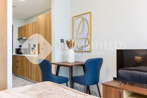 Een moderne hotelkamer met een bed, een televisie, een bureau met twee stoelen, een kitchenette en een kunstwerk in een lijst aan de muur.