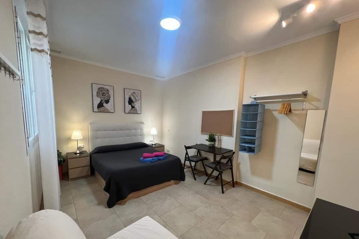 A bedroom with a bed, bedside table, lamp, and a desk with a chair. There is a mirror on the wall and a shelf with towels.