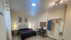 Un dormitorio con cama, mesita de noche, lámpara y escritorio con silla. Hay un espejo en la pared y un estante con toallas.
