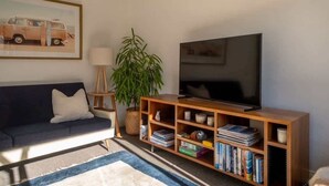 A living room with a dark sofa, a wooden TV stand with books, a TV, a lamp, and a plant.