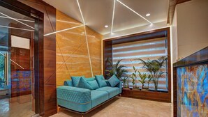 A modern living room with a blue sofa, wooden accents, and a large window with plants. - SRS Suite (Bengaluru)