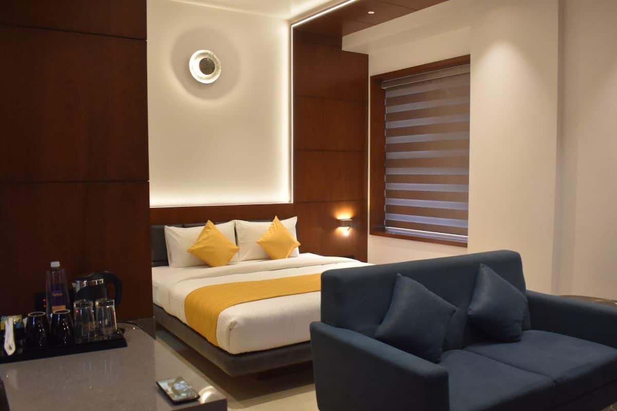 A modern hotel room with a bed, a blue sofa, a bedside table with a phone, and a window with blinds.