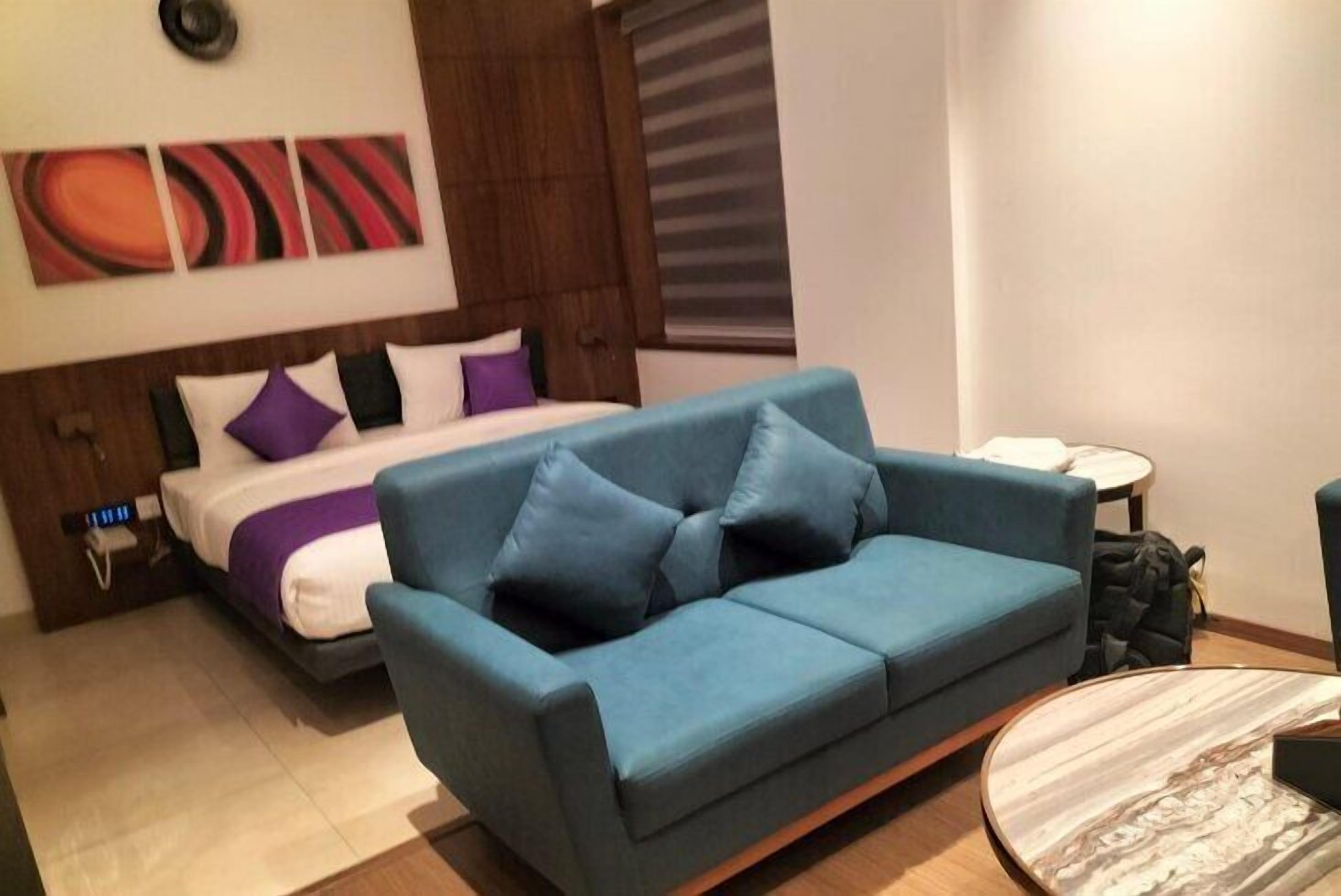 A modern hotel room with a bed, a blue sofa, a round coffee table, and abstract wall art.
