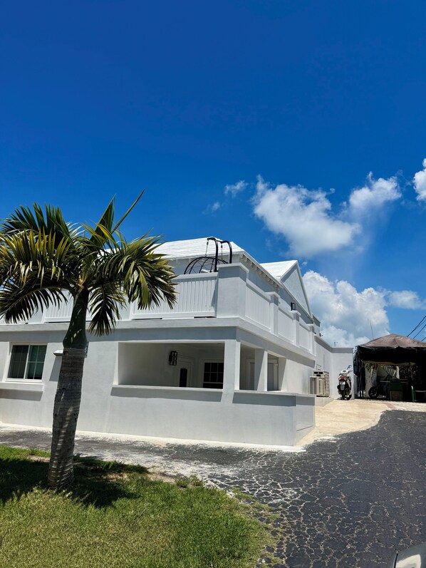 A two-story white building with a balcony, a palm tree in the foreground, and a clear blue sky.