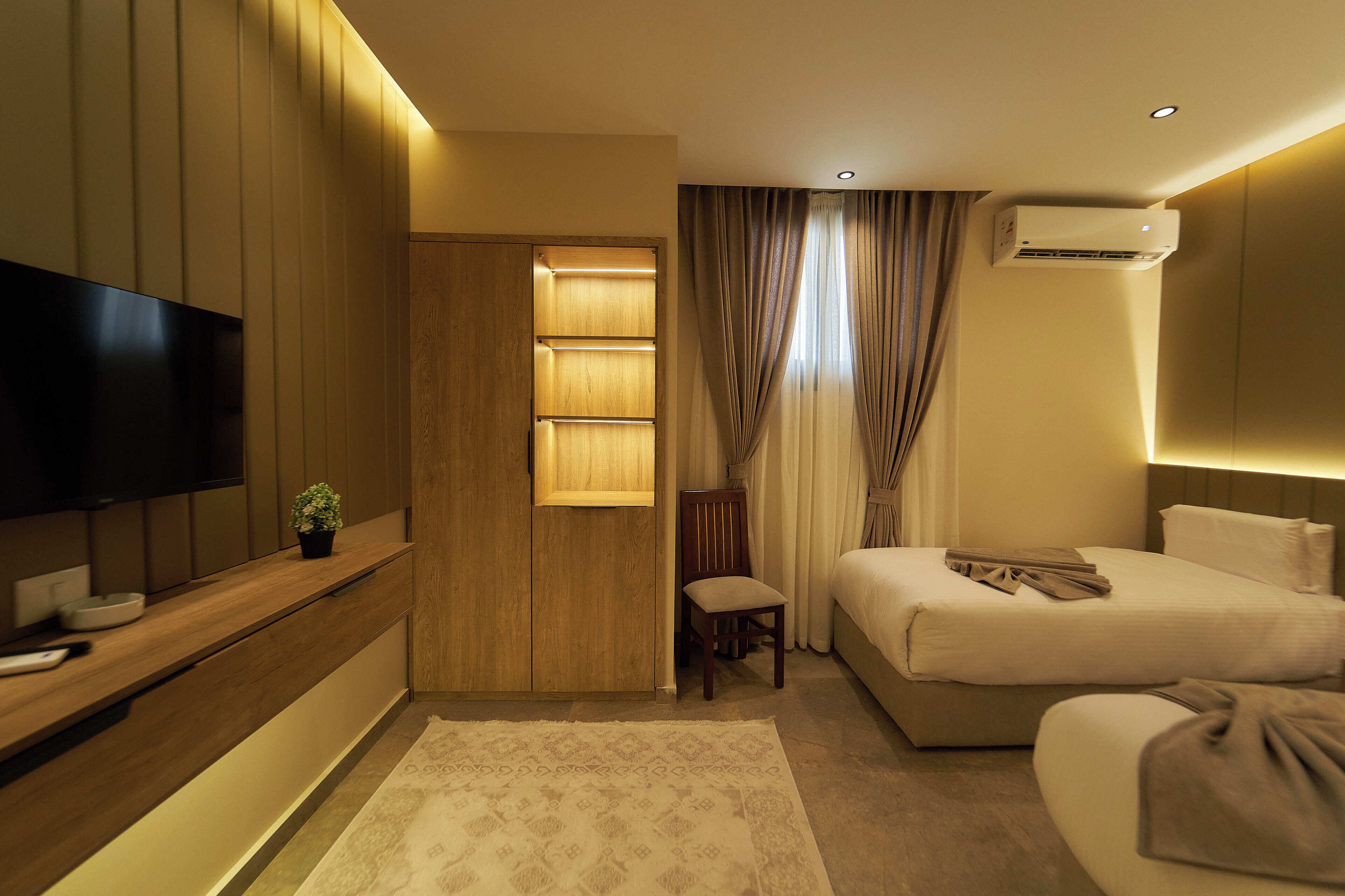 A modern hotel room with a flat-screen TV, a wooden wardrobe, a bed, a chair, and an air conditioning unit.