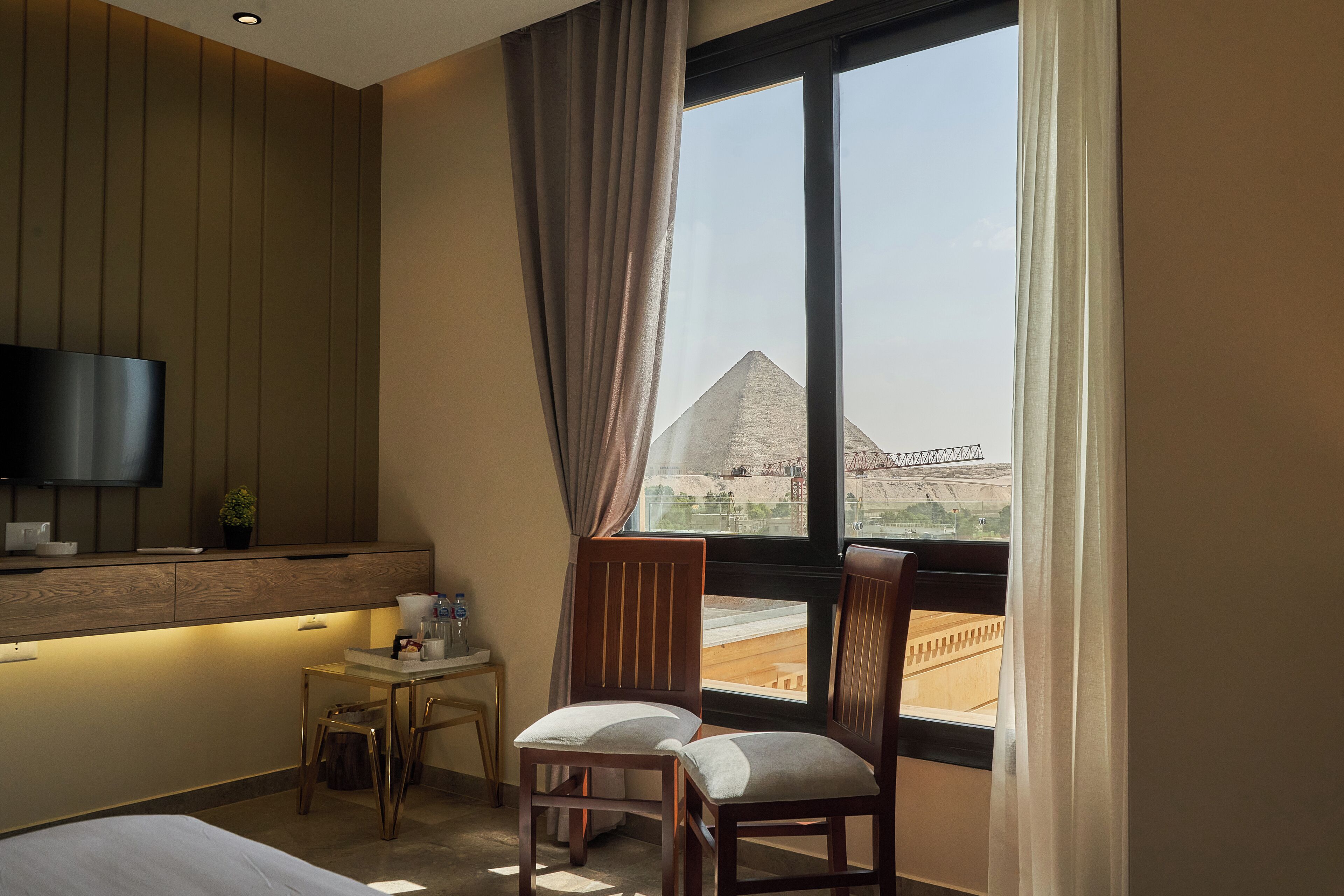 Habitación de hotel con cama, escritorio, silla, televisión y ventana con vista a una pirámide.