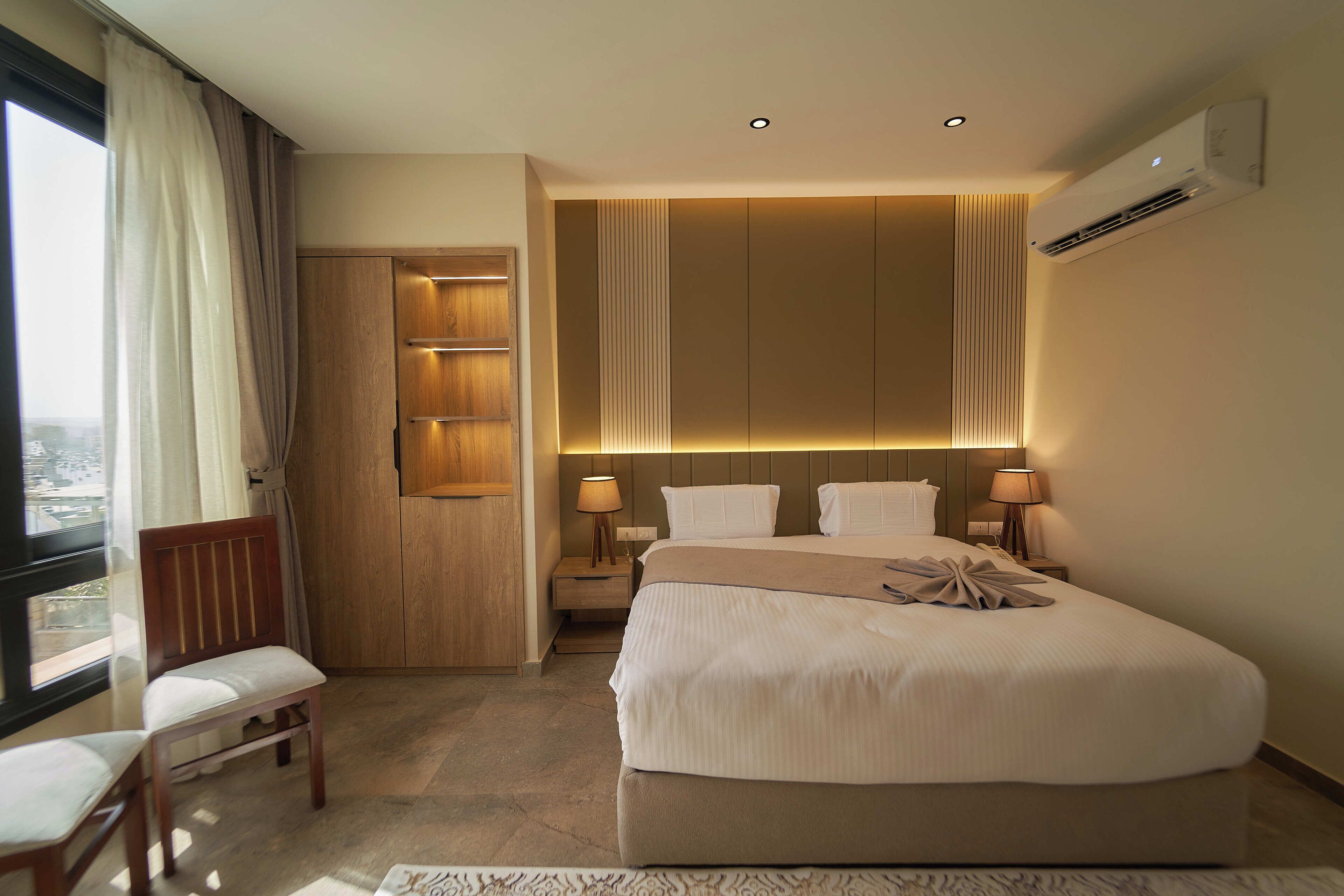 Ein Hotelzimmer mit einem großen Bett, einem Holzschrank, einem Nachttisch mit Lampen und Blick durch ein Fenster.