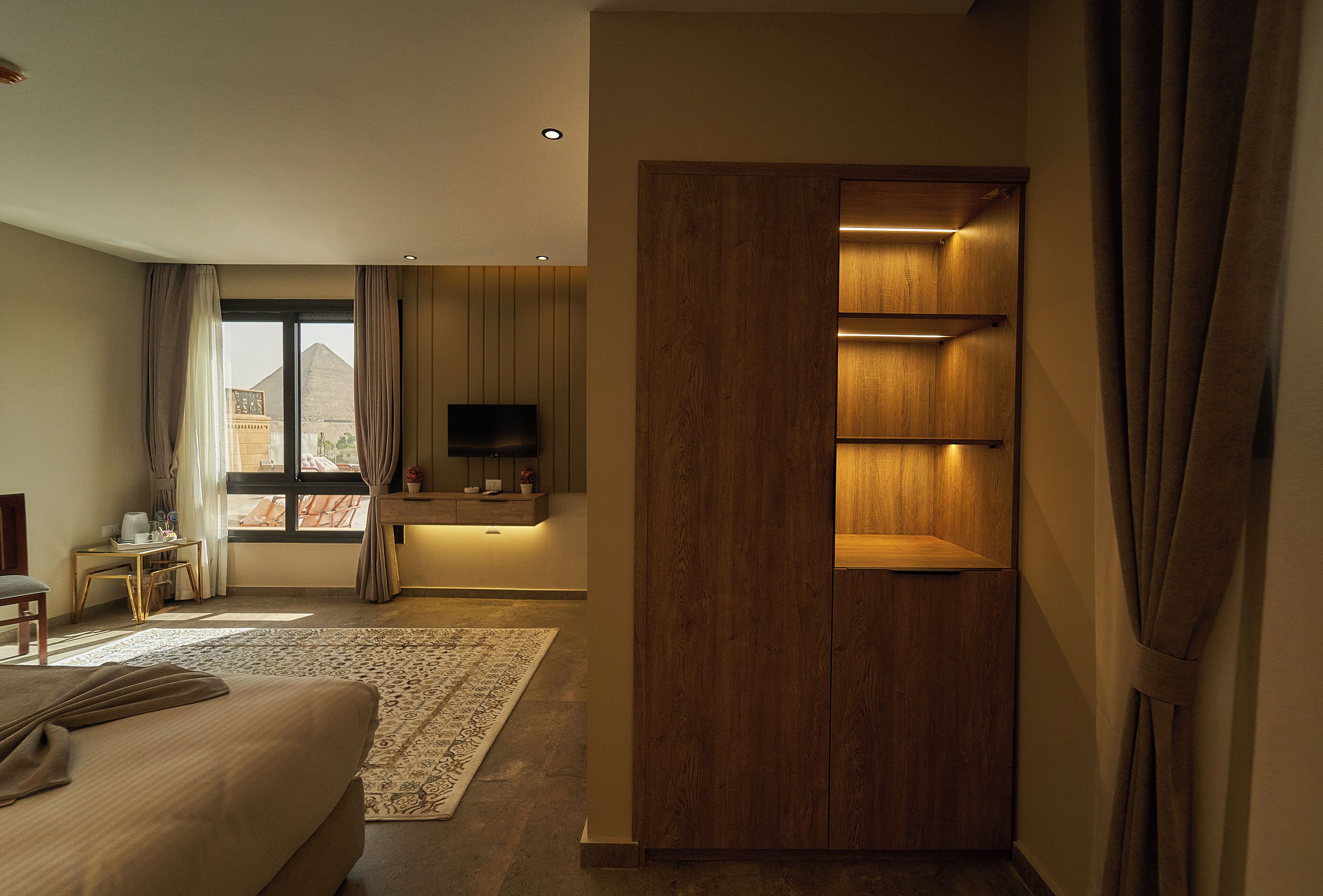 Une chambre d’hôtel moderne dotée d’un grand lit, d’un bureau avec un téléviseur à écran plat, d’une armoire en bois et offrant une vue sur une pyramide par la fenêtre.