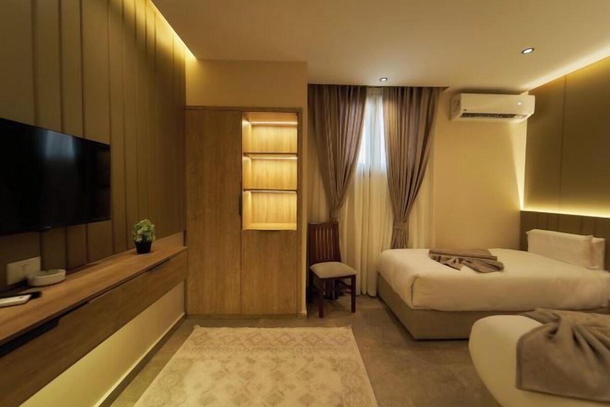 A modern hotel room with a large bed, a smaller bed, a TV, a desk with a plant, and a wardrobe.