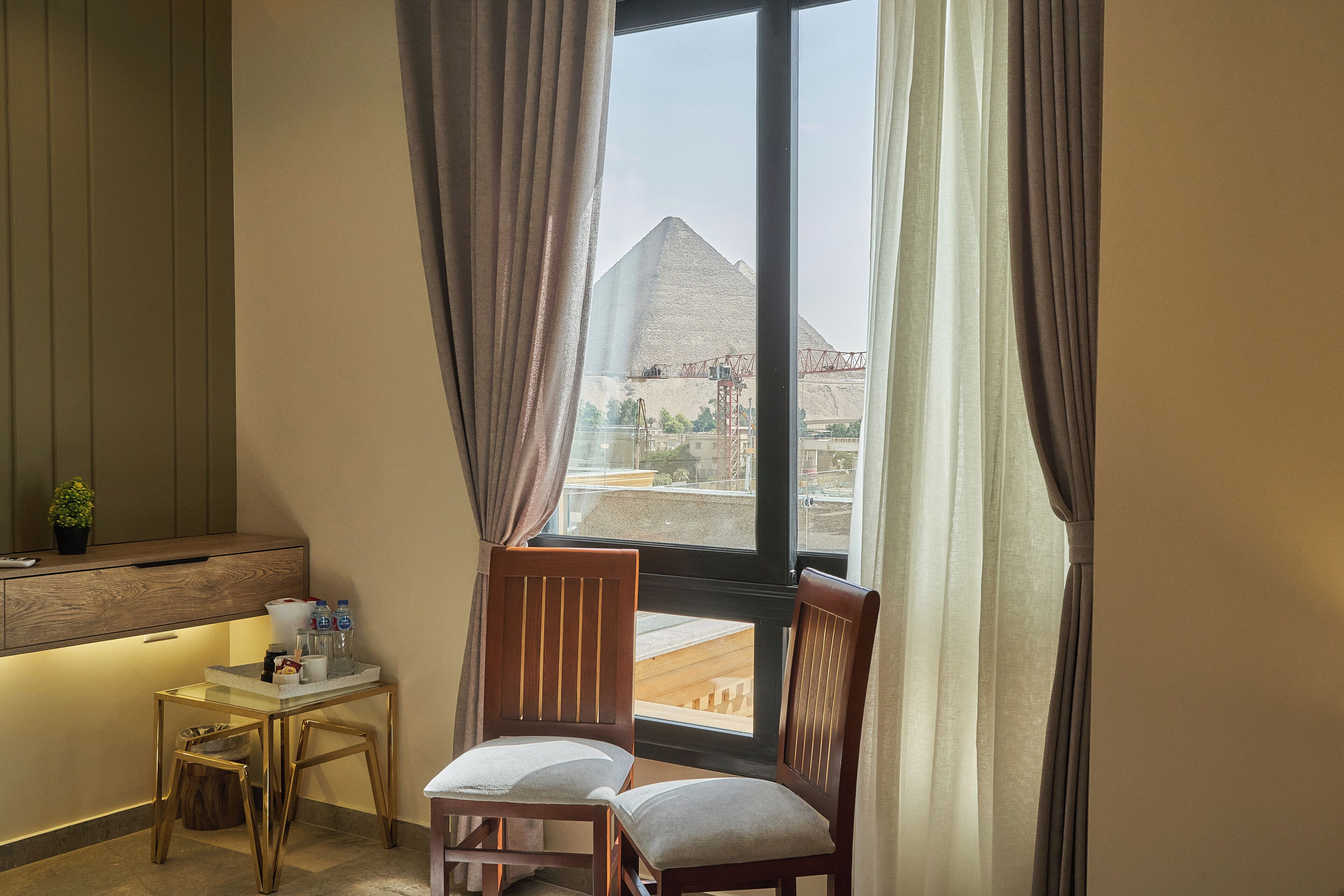 A hotel room with a desk, two chairs, a small table, and a view of a pyramid through the window.