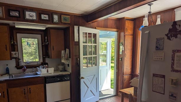 A kitchen with wooden cabinets, a white dishwasher, and a refrigerator. There is a window with a view of greenery outside.