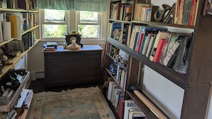 A room with a large bookshelf filled with books, a desk with a clock, and a window with curtains.