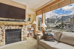 A cozy living room with a stone fireplace, a bookshelf, a comfortable sofa, and a view of a snowy mountain. - Tx204 Taylors Crossing 3 Bedroom Condo (Copper Mountain)