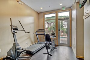 Une salle de sport équipée d’appareils de cardio et d’un vélo d’appartement, une grande fenêtre donnant sur des immeubles et une piscine à l’extérieur.