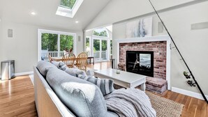 A spacious living room with a fireplace, a dining area, and large windows offering a view of the outdoors. - Hyannis Hideaway: Pool, Beaches & Main Street Fun RPAM114 (Hyannis)