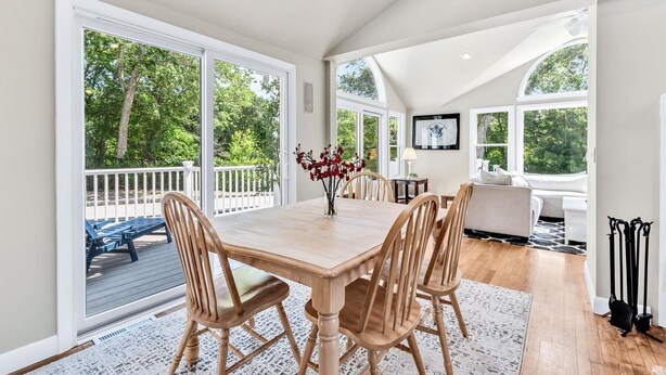 A spacious dining area with a large wooden table and chairs, large windows offering views of trees, and a cozy seating area with a sofa and lamp. - Hyannis Hideaway: Pool, Beaches & Main Street Fun RPAM114 (Hyannis)