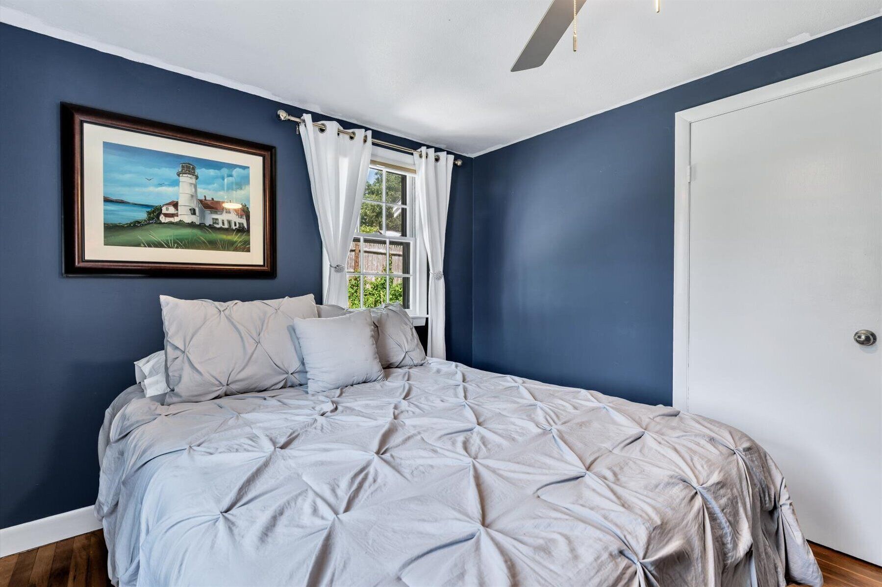 A bedroom with a large bed, a framed picture of a lighthouse, and a window with white curtains.