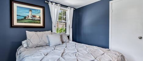 A bedroom with a large bed, a framed picture of a lighthouse, and a window with white curtains.