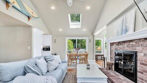 A spacious living room with a sofa, coffee table, and fireplace. There is a dining area with a table and chairs, and a kitchen area with cabinets and a microwave. - Hyannis Hideaway: Pool, Beaches & Main Street Fun RPAM114 (Hyannis)