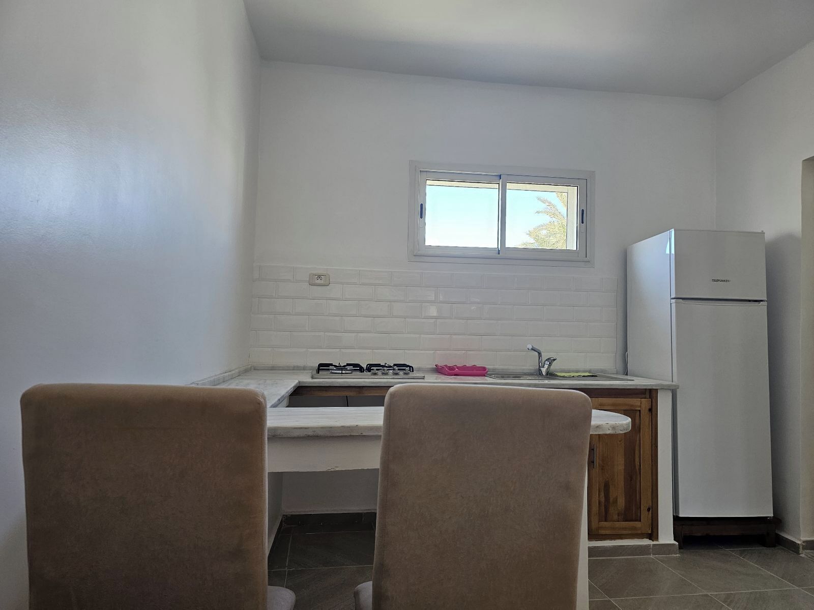 Une cuisine compacte avec des murs en briques blanches, une fenêtre, une cuisinière à gaz, un évier et un réfrigérateur.