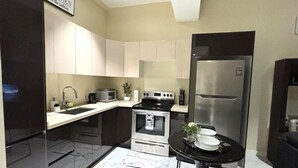 A compact kitchen with white cabinets, a black refrigerator, a stove, and a small dining table with a black chair. - One Bedroom City Apartment with Pool (Kingston)