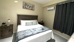 A bedroom with a bed, a nightstand, a lamp, a wall-mounted air conditioner, and a painting. - One Bedroom City Apartment with Pool (Kingston)