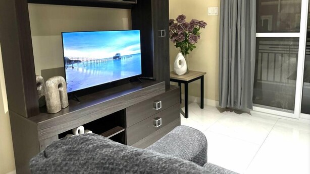 A living room with a flat-screen TV on a dark wooden entertainment unit, a small table with a vase of flowers, and a grey sofa. - One Bedroom City Apartment with Pool (Kingston)