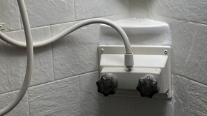 Une salle de bain avec des murs carrelés de carreaux blancs et des toilettes blanches dont la tuyauterie est visible.