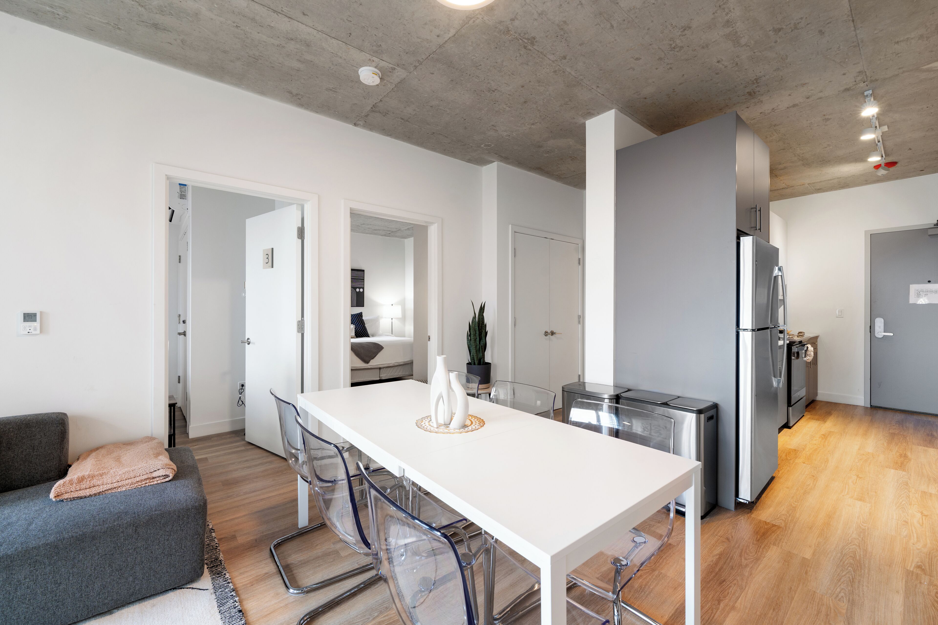 Una cocina moderna con encimera blanca, electrodomésticos de acero inoxidable y un comedor con mesa blanca y sillas transparentes.