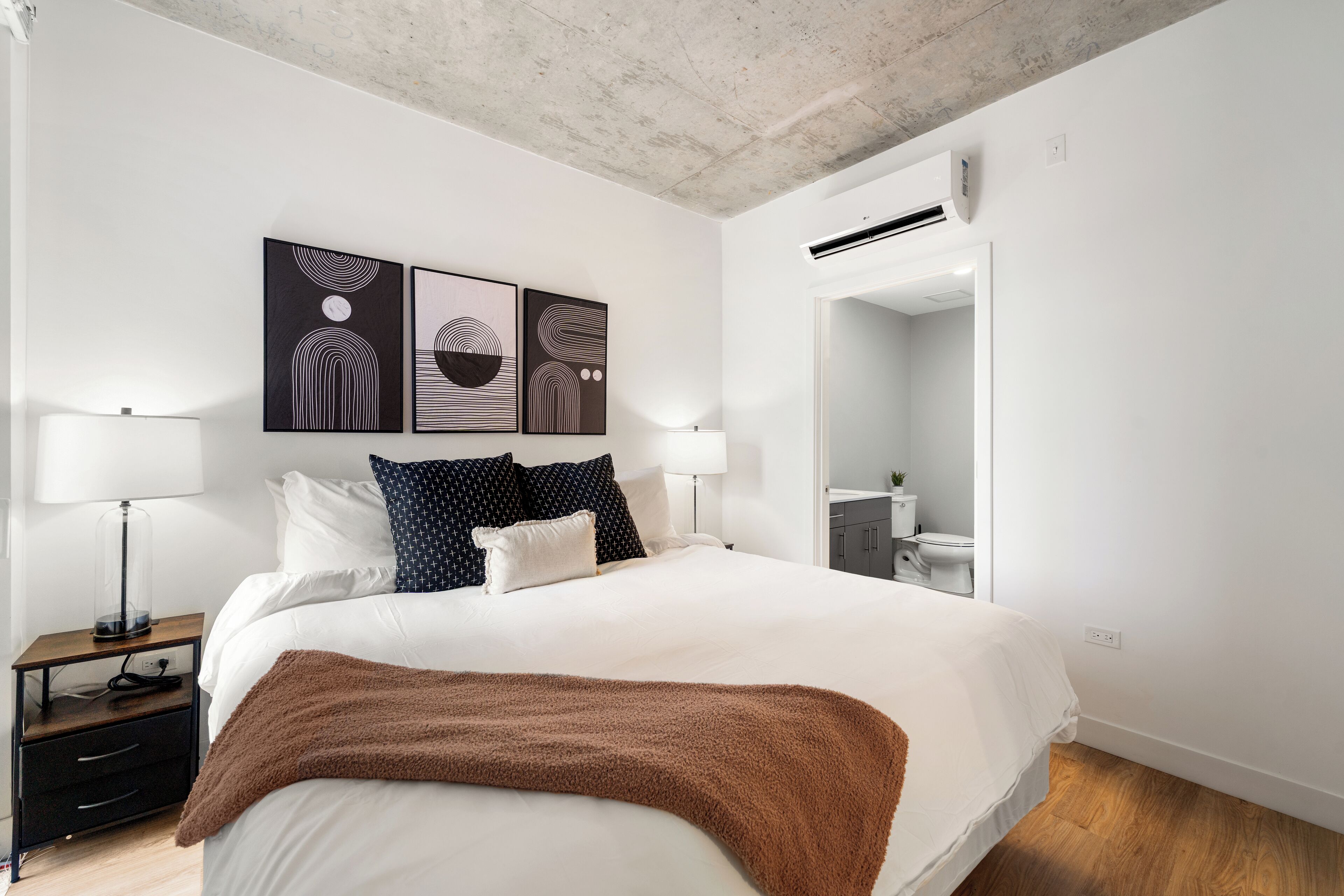 Une chambre à coucher moderne avec un grand lit, deux tables de chevet, un plaid marron et des œuvres d’art abstraites accrochées au mur.