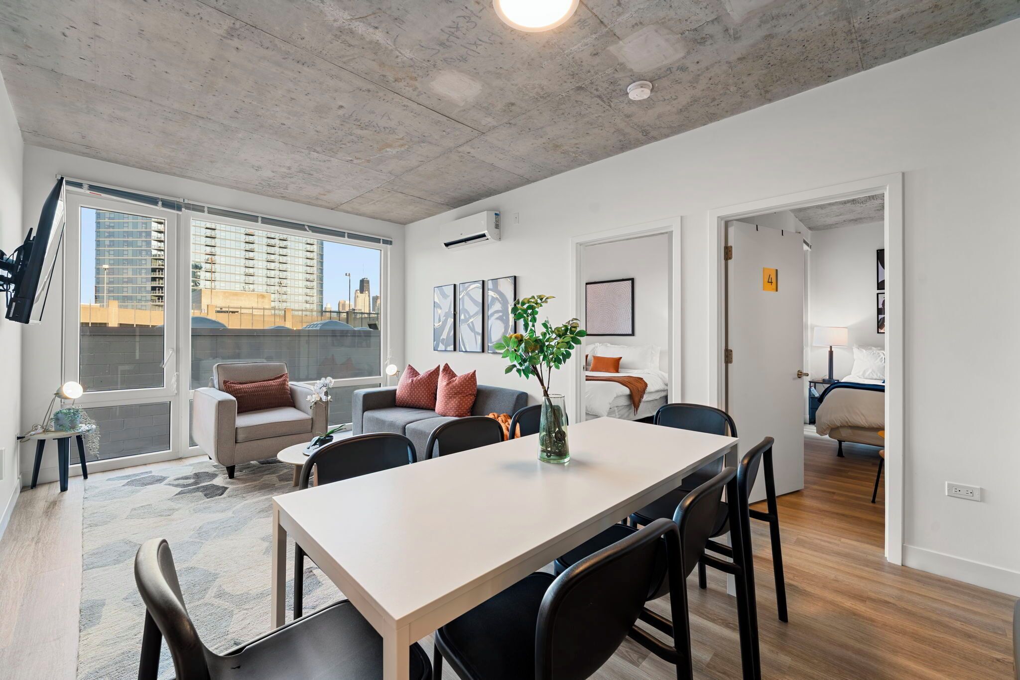 A modern apartment with a dining area, a living room with a sofa and a coffee table, and a bedroom visible through an open door.