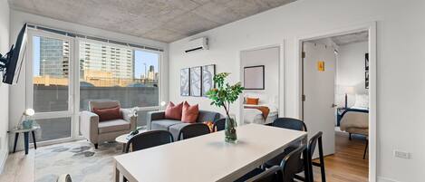 A modern apartment with a dining area, a living room with a sofa and a coffee table, and a bedroom visible through an open door.