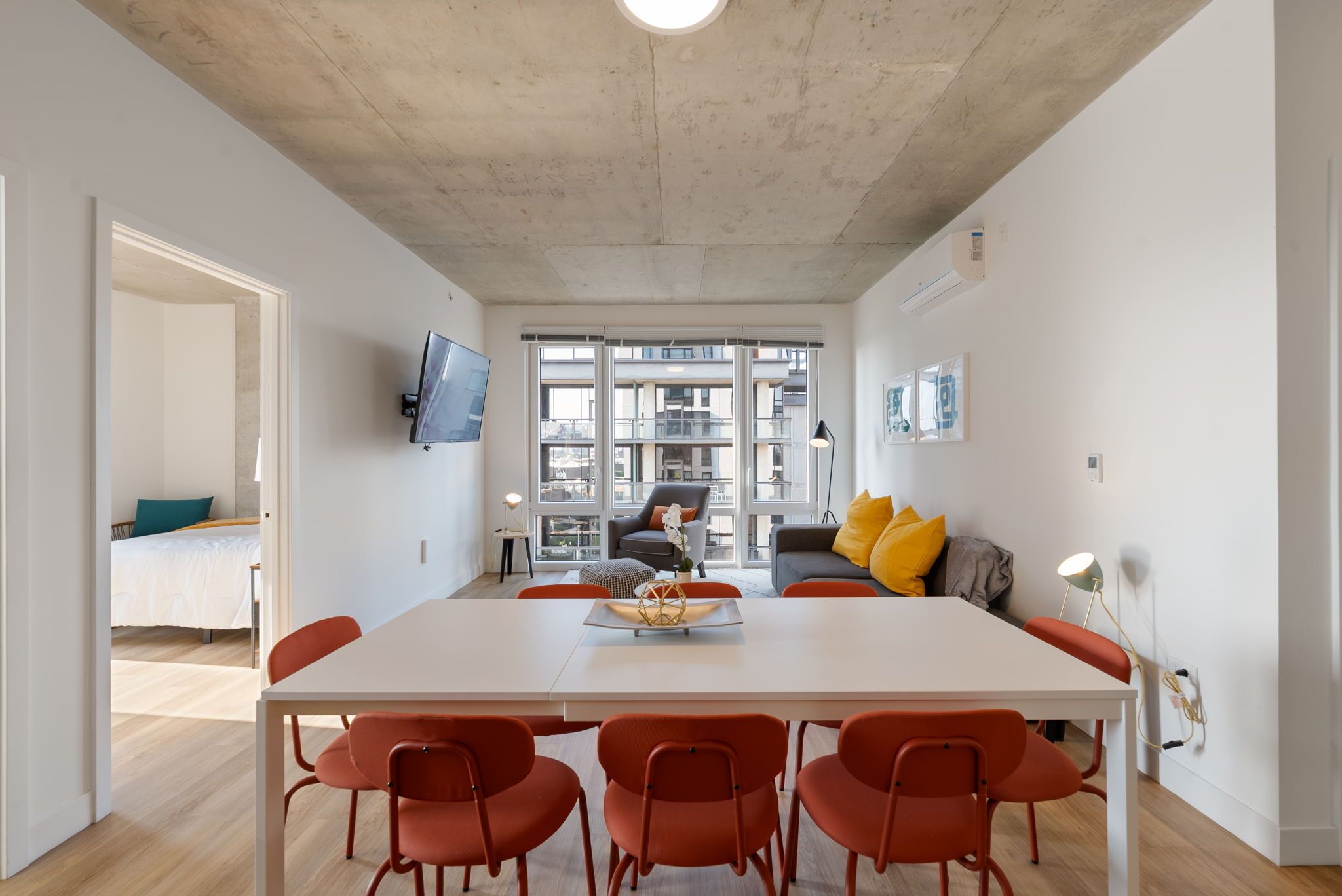 A modern living room with a white dining table, orange chairs, a sofa with yellow pillows, and a TV mounted on the wall.