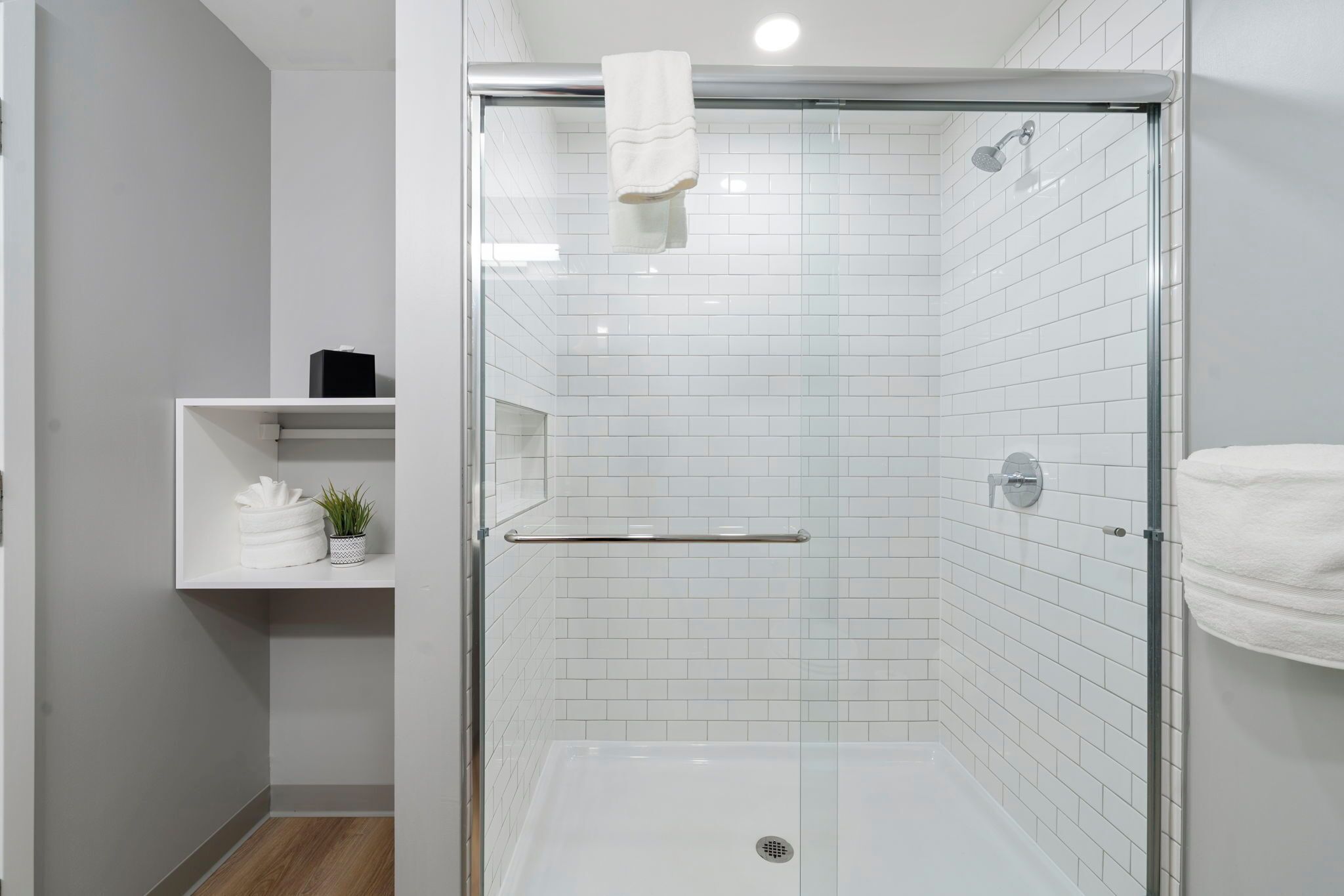 A modern bathroom with a glass-enclosed shower, white tiled walls, and a shelf with towels and a plant.