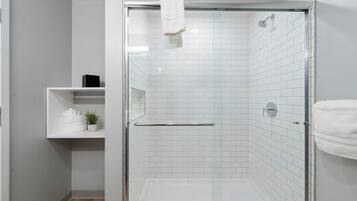 A modern bathroom with a glass-enclosed shower, white tiled walls, and a shelf with towels and a plant.